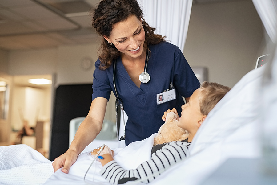 Beautiful kind female nurse taking care of little boy hospitalized in bed. Happy woman nurse tuck the covers back to the young child patient lying on hospital bed. Friendly medical worker talking with kid patient in pediatrics ward at clinic.