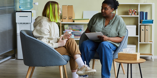 African female social worker talking to difficult teenage girl while they sitting on armchairs at office
